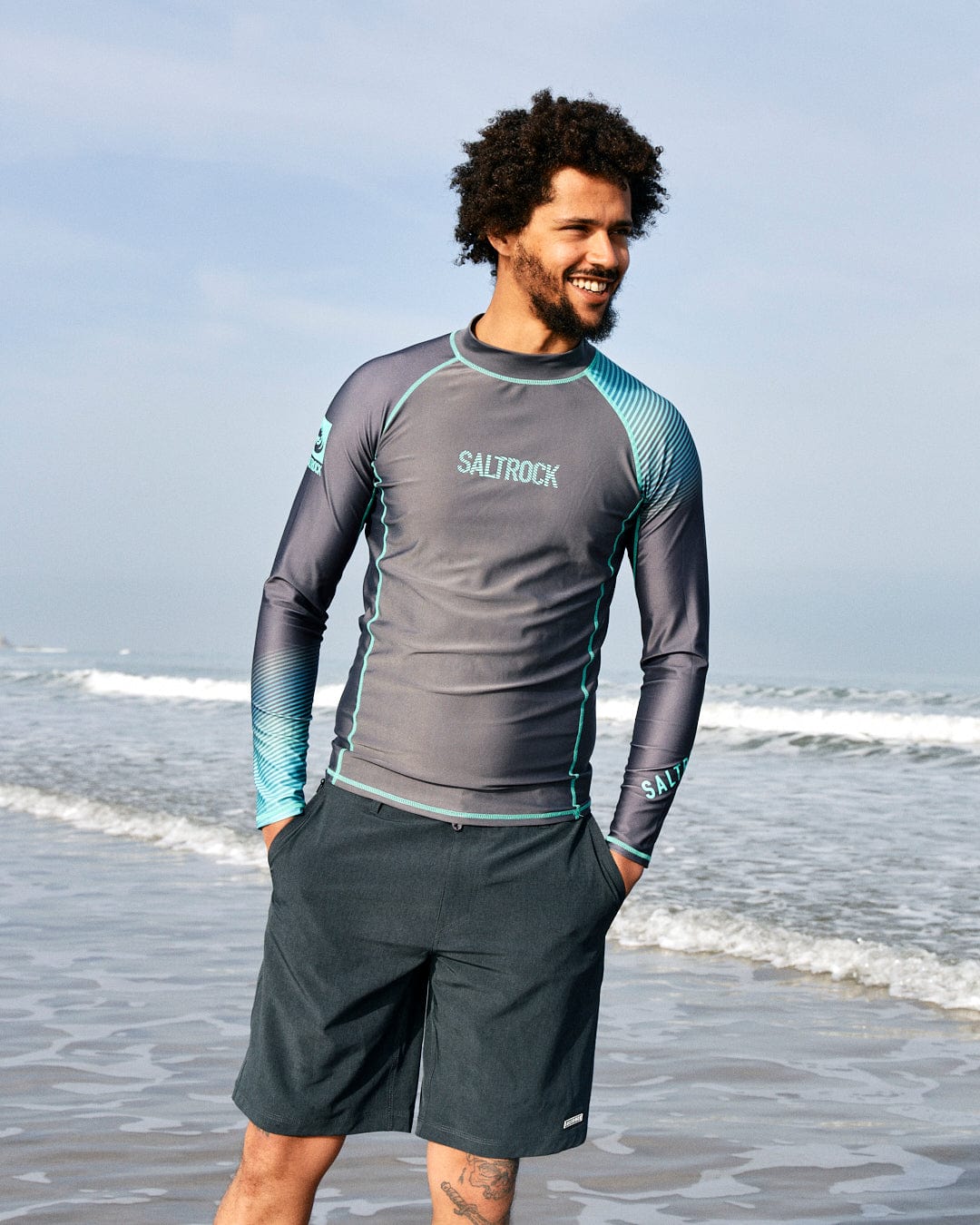 Man with curly hair smiling at the beach, wearing a gray and teal Saltrock DNA Wave - Mens Long Sleeve Rashvest - Grey/Turquoise and black shorts.