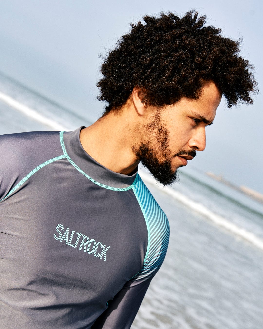 A man with curly hair wearing a Saltrock DNA Wave - Mens Long Sleeve Rashvest in Grey/Turquoise stands on a beach, looking off to the side with a focused expression.
