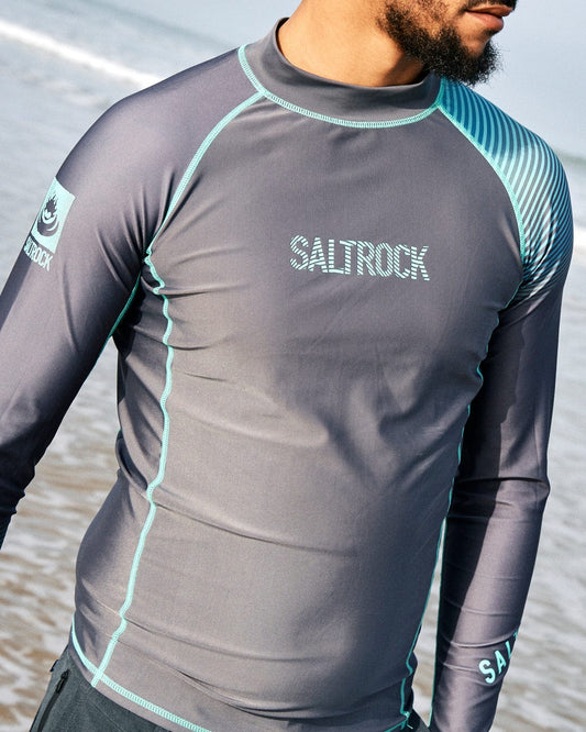 Man wearing a gray Saltrock DNA Wave - Mens Long Sleeve Rashvest with turquoise accents, standing on a beach; only his torso and one arm are visible.