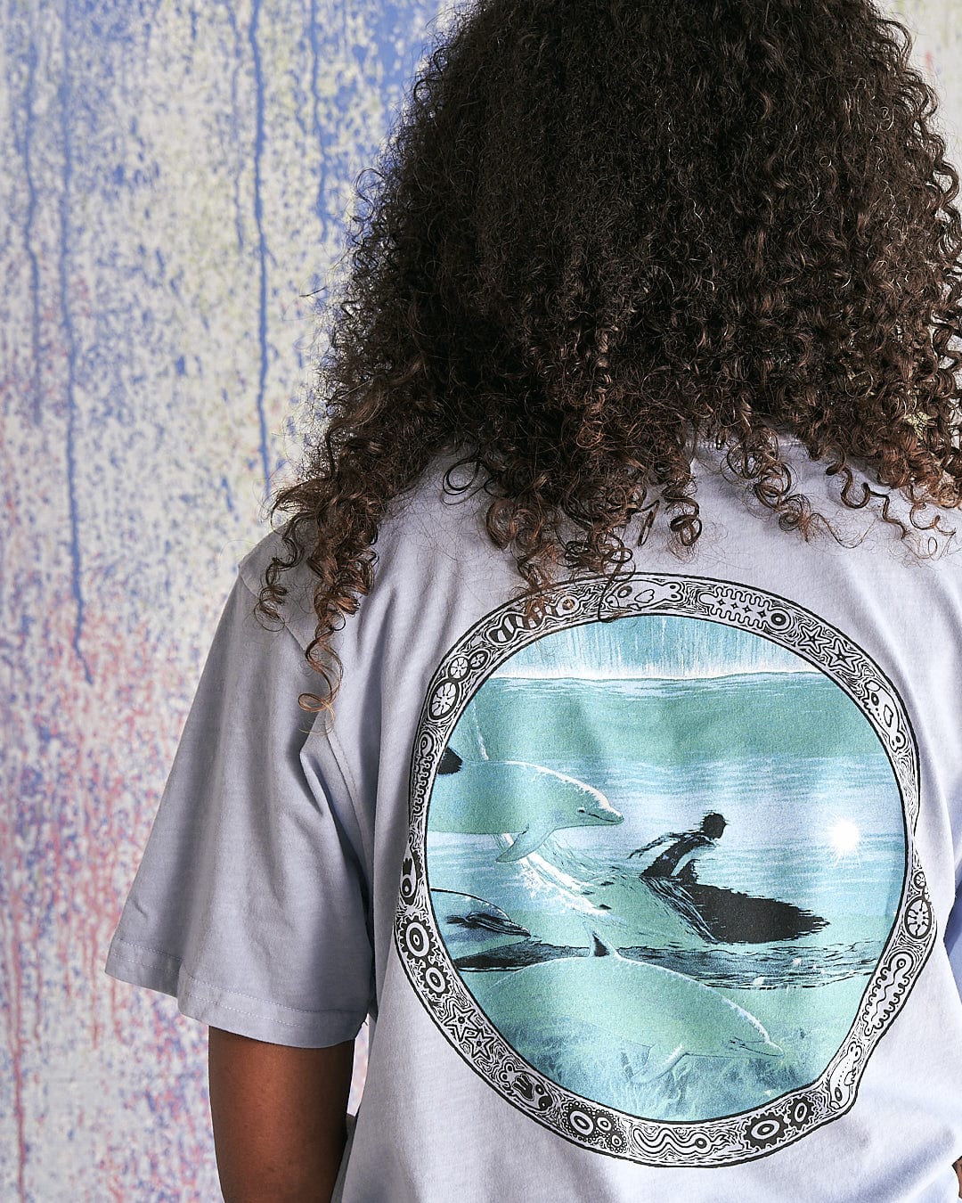 The back of a woman wearing a Saltrock Dolphin - Limited Edition 35 Years T-Shirt with an image of a woman riding a surfboard.