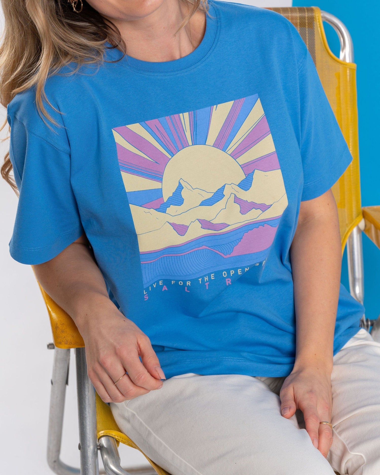 A person relaxes in a large yellow chair, wearing the Saltrock Open Spaces oversized short sleeve t-shirt in blue, featuring a vivid mountains & sun design.