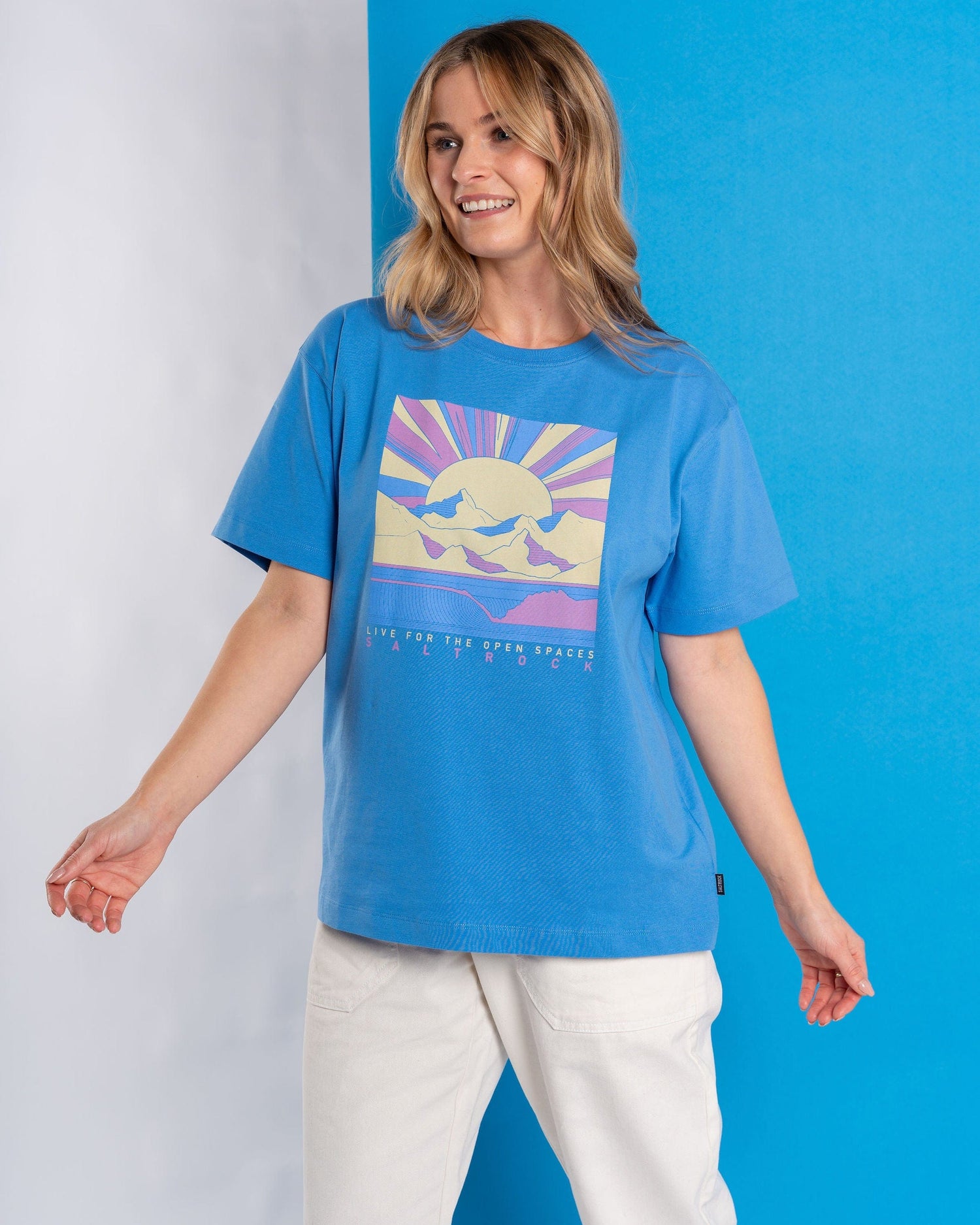 A person stands against a blue background, wearing Saltrock's Open Spaces Women's Oversized Short Sleeve T-Shirt in blue, featuring a vivid mountains & sun graphic, perfectly paired with white pants.