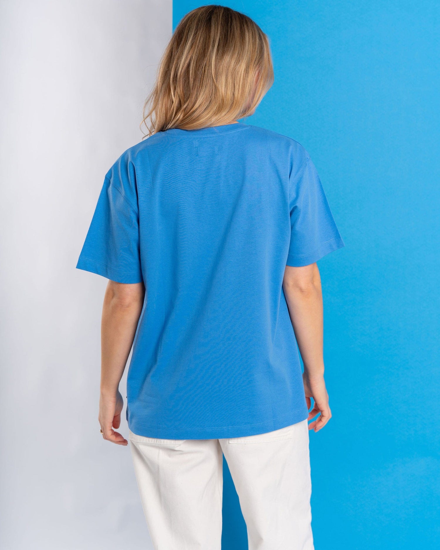 A long-haired person wears a Saltrock "Open Spaces" women's oversized blue T-shirt and white pants, standing against a mountains and sun graphic in blue and white, facing away.