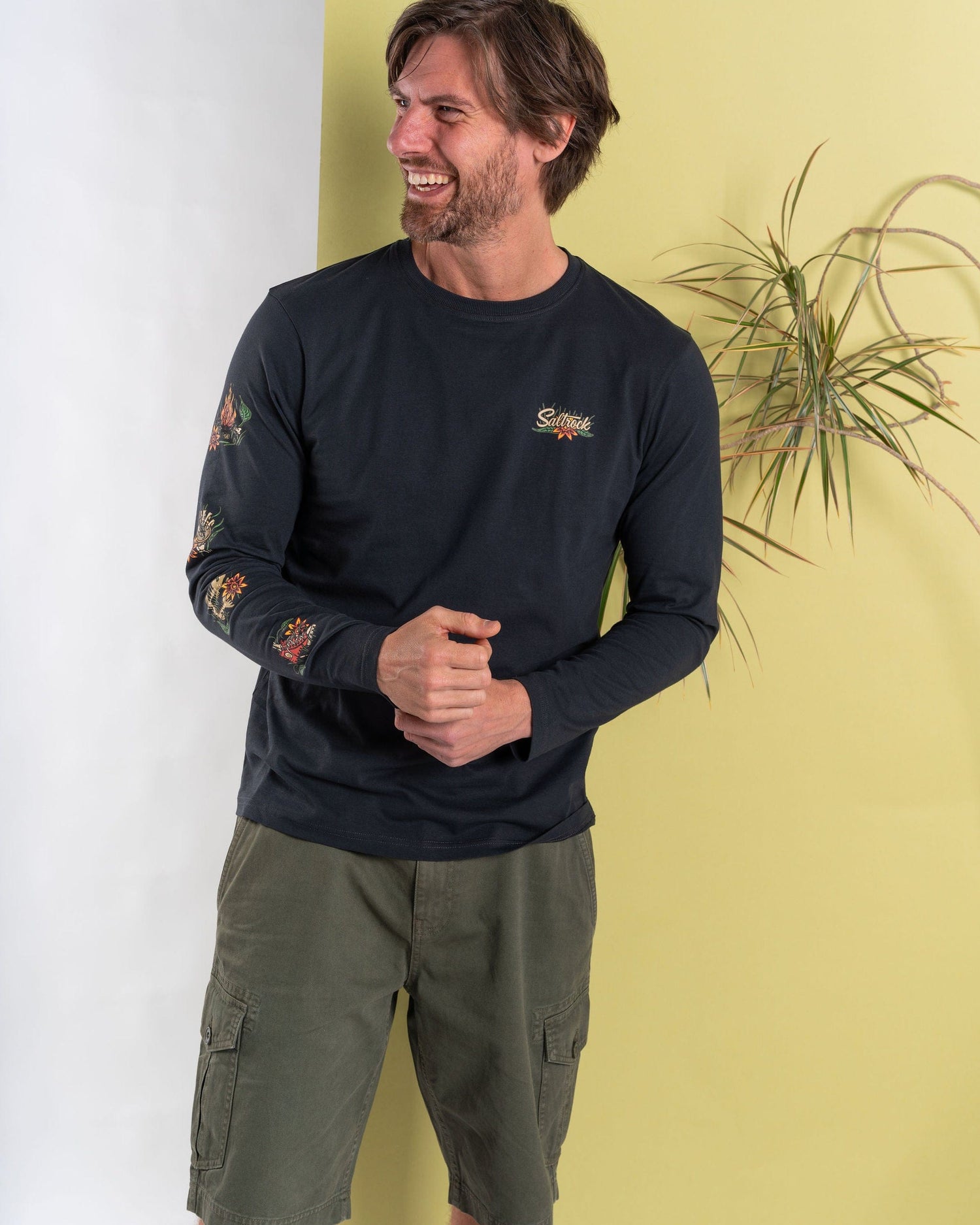 A man stands laughing against a vibrant yellow and white backdrop framed by a plant, wearing the Saltrock Lantern Men's Long Sleeve T-Shirt in dark grey and green shorts, which capture the brand's adventurous spirit with its branded graphics.