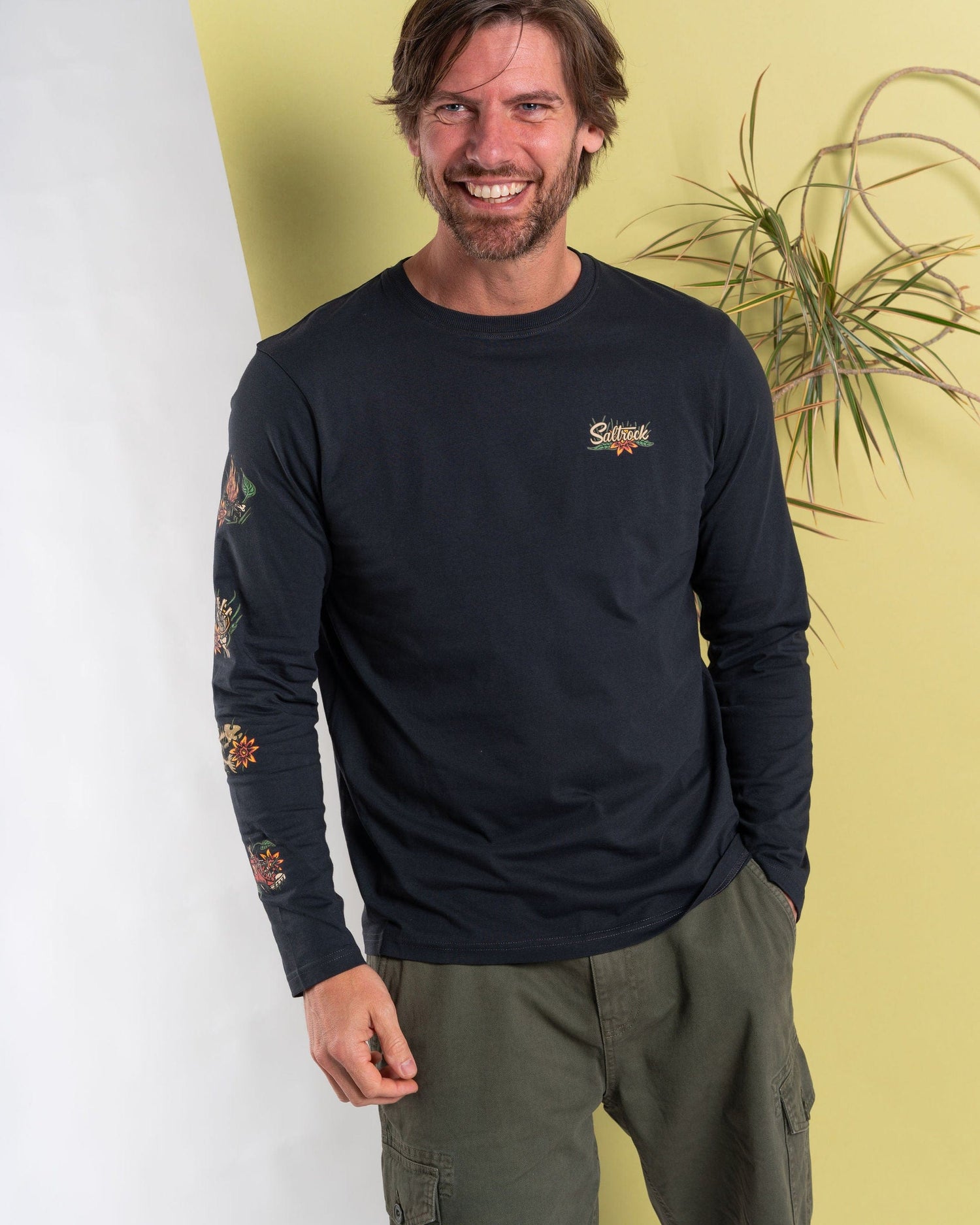 A man in a Saltrock Lantern dark grey long-sleeve T-shirt with floral embroidery, made of 100% cotton, complements it with green cargo pants as he stands by a plant against a bright yellow and white backdrop, embodying Saltrock's laid-back style.