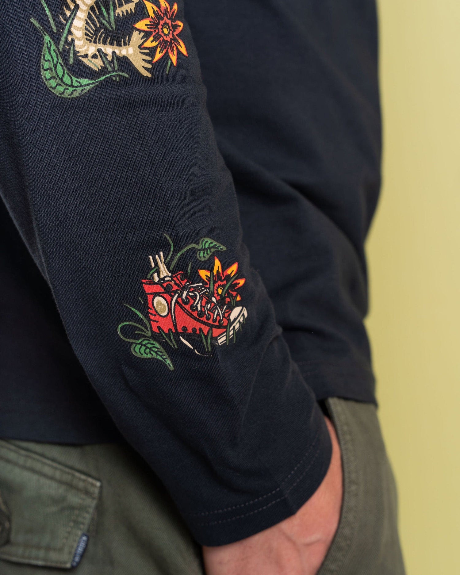 Close-up of the Saltrock Lantern men's long sleeve t-shirt in dark grey, crafted from 100% cotton. It features colorful abstract floral and geometric designs on the arm. Paired with green pants, the vibrant graphics stand out against a light yellow background.