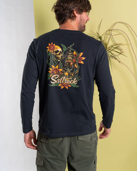 A man in Saltrock's dark grey Lantern long-sleeve T-shirt with branded graphics on the back and green cargo pants stands confidently against a split green and white background, next to a plant.