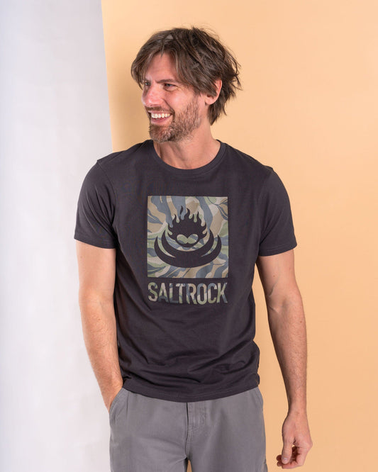 A man with medium-length hair and a beard beams while wearing a Saltrock Camo Leaf Flame men's short sleeve t-shirt in dark grey, featuring a camo print logo, set against a split beige and white background.