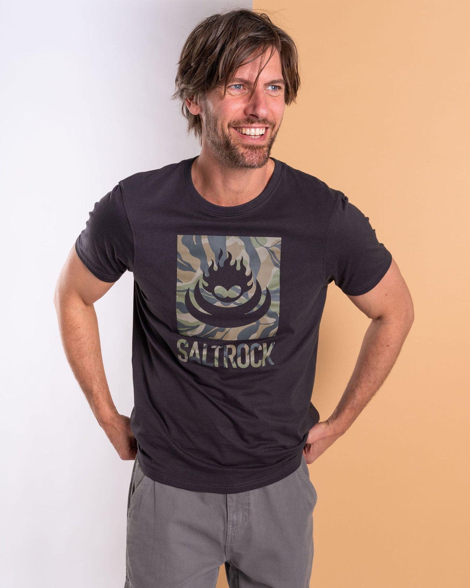 A man in a dark grey Saltrock "Camo Leaf Flame" t-shirt, made from 100% cotton, stands against a split white and beige background. With hands on hips, he smiles confidently and exudes a relaxed vibe.