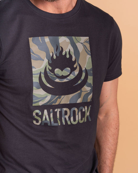 A person wears the Camo Leaf Flame Saltrock tee in dark grey, boasting a striking camo print. Made from 100% cotton, it combines comfort and style with an iconic logo.