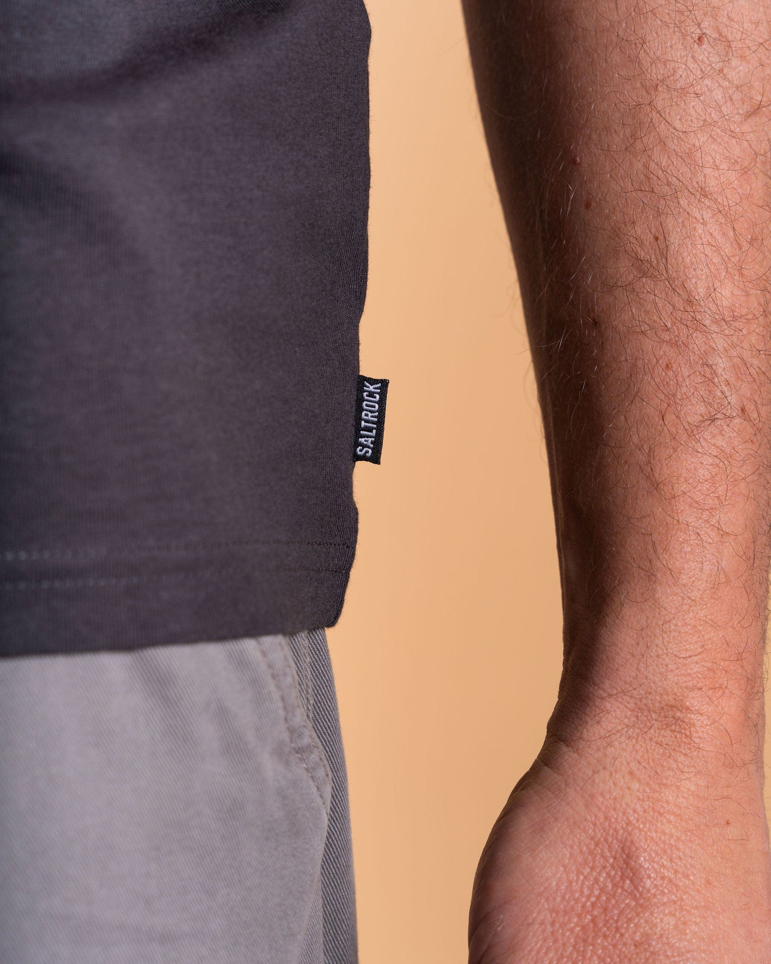 Close-up shot of a person wearing the Saltrock Camo Leaf Flame Men's Short Sleeve T-Shirt in dark grey, paired with light gray pants, against a beige background.