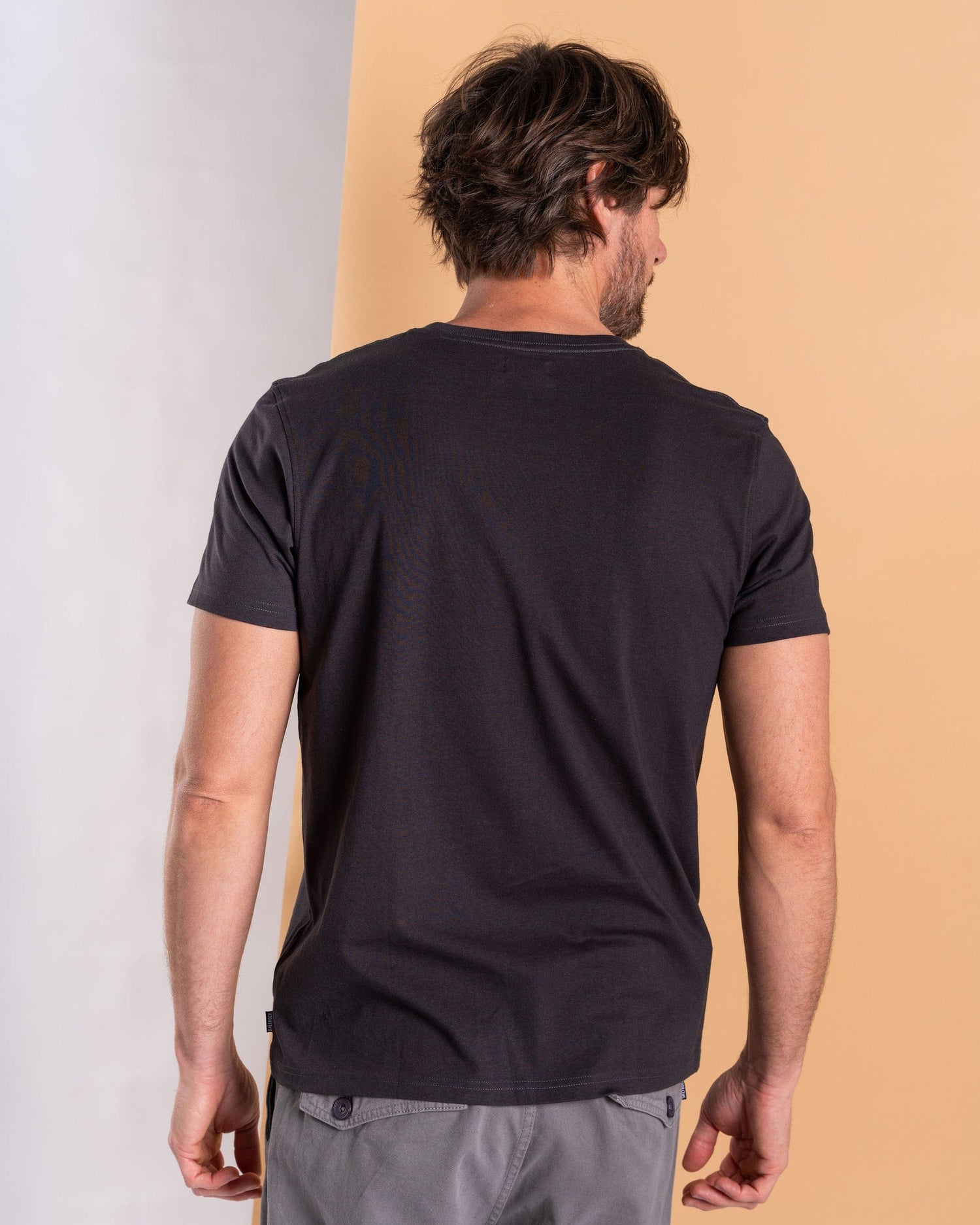 A dark-haired man, facing away from the camera, wears a Saltrock Camo Leaf Flame Dark Grey T-shirt and gray pants in front of a two-tone background.