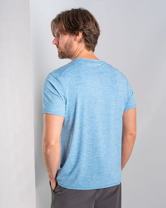 A man with brown hair and a beard stands with his back turned, wearing the Saltrock Aestus Men's UPF 50+ Hybrid T-Shirt in blue along with gray pants made from recycled materials, in a plain indoor setting.
