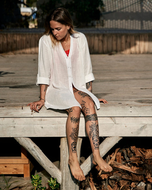 Woman sitting on a wooden dock, wearing a Saltrock Karthi Womens Long Sleeve Shirt in White and shorts, with visible leg tattoos, looking thoughtfully to the side.