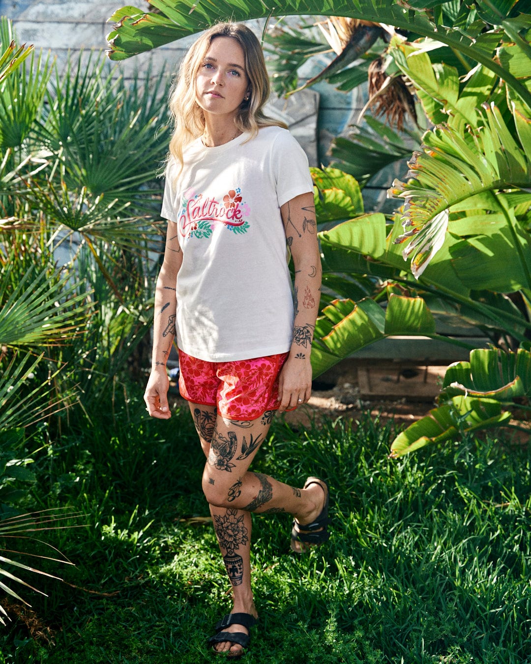 Woman with tattoos standing in a garden, wearing a white t-shirt and Saltrock Hibiscus Womens Boardshorts in Red/Pink with an elasticated drawstring waist, looking at the camera.