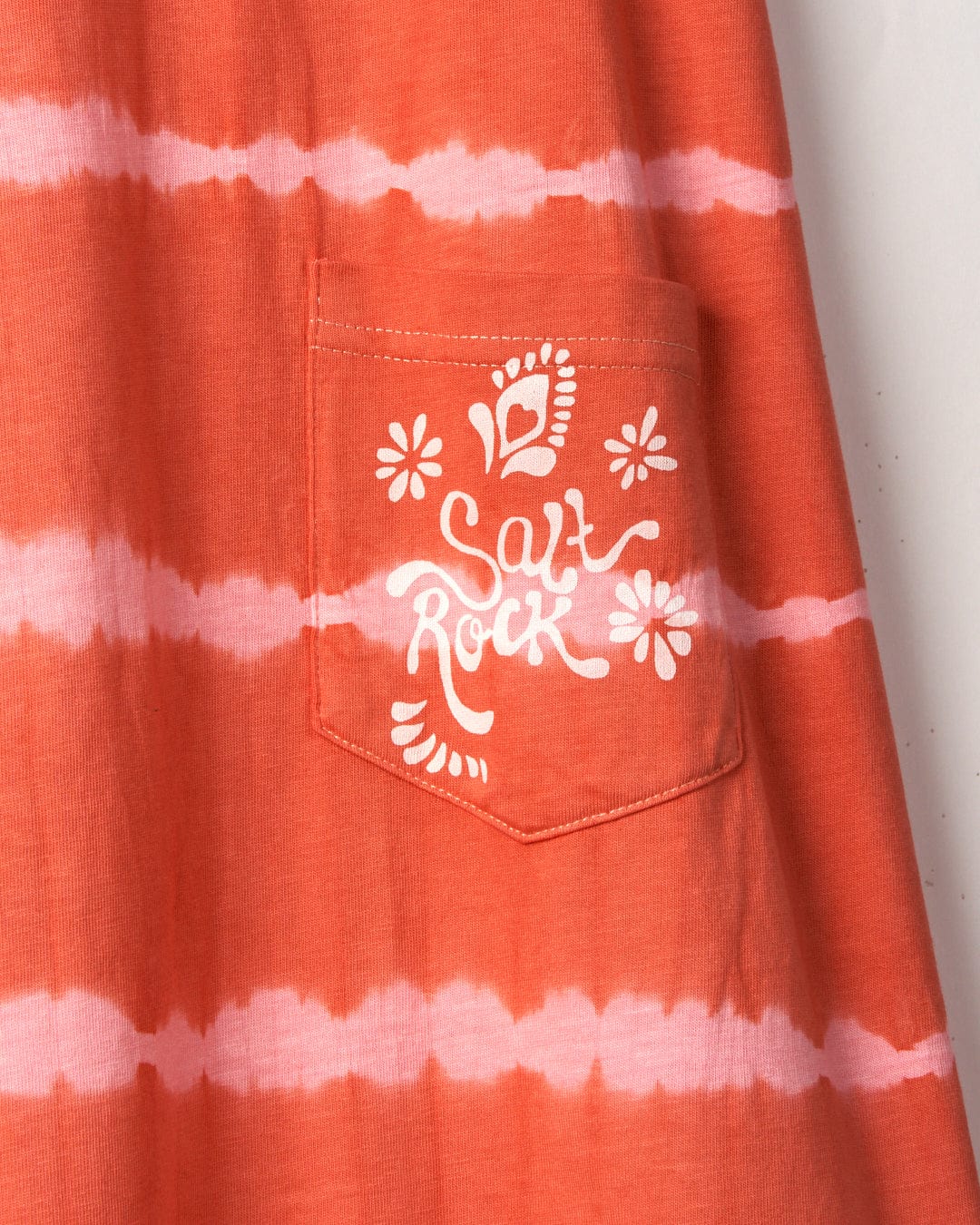 An orange and white tie-dye print fabric on the Eliana - Kids Tie Dye Vest Dress - Peach features a pocket with the phrase "Saltrock" and floral designs printed in white.