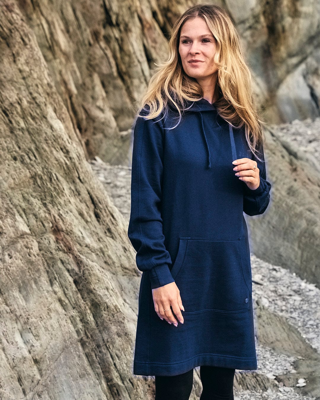 A woman wearing a Elsa - Womens Hooded Sweat Dress - Dark Blue from Saltrock, standing on a rock, embodying style and comfort.