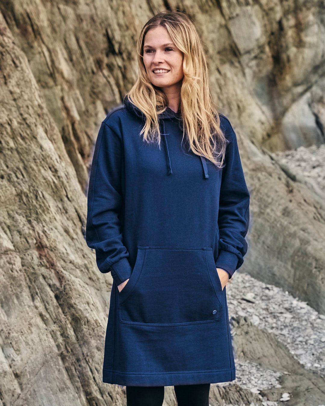 A woman in a Saltrock Elsa - Womens Hooded Sweat Dress - Dark Blue standing on a rock, showcasing both comfort and style.