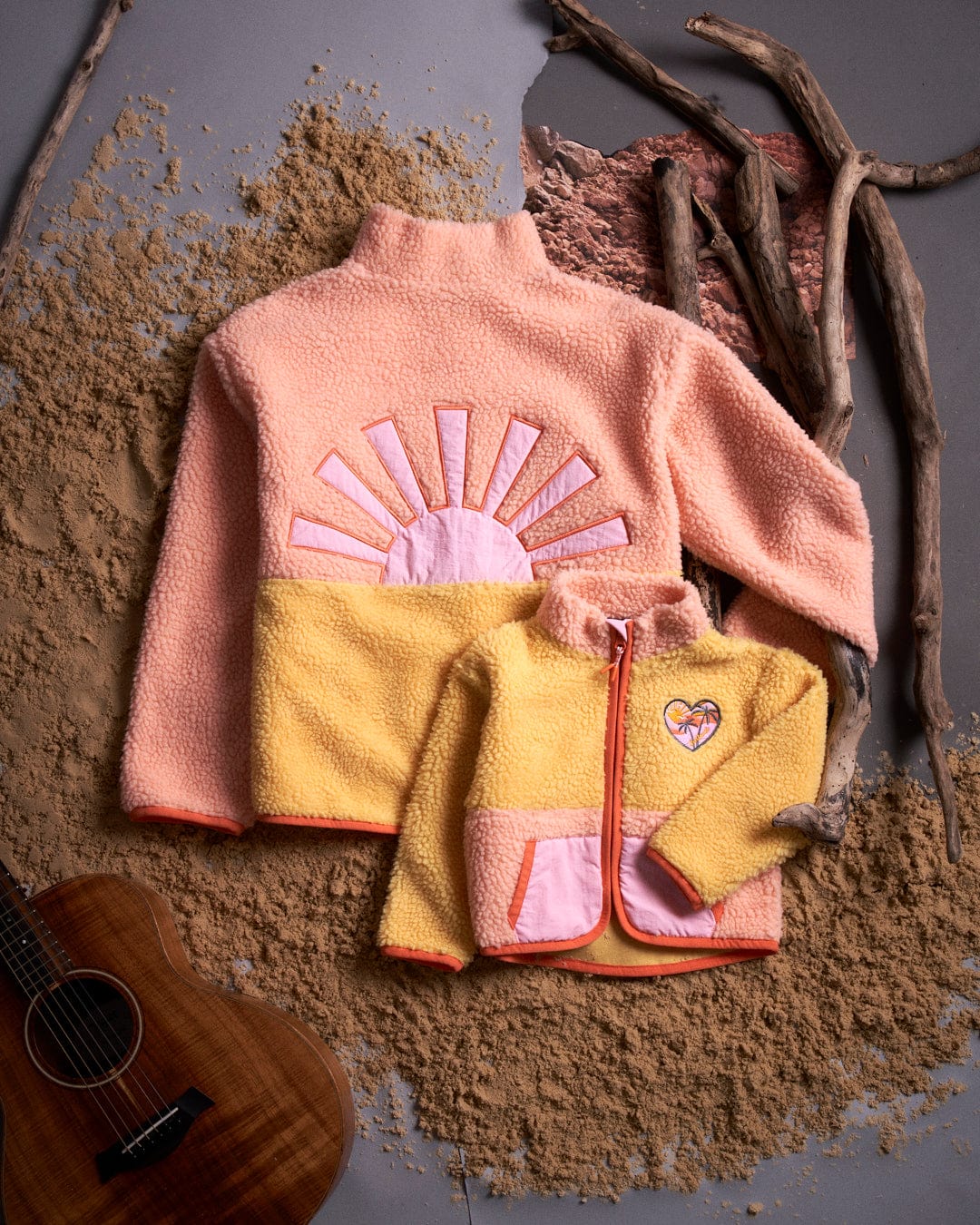 A girl wearing a Saltrock jacket and an Emery Sunshine Kids Sherpa Fleece - Yellow embroidered sunrise guitar on sand.