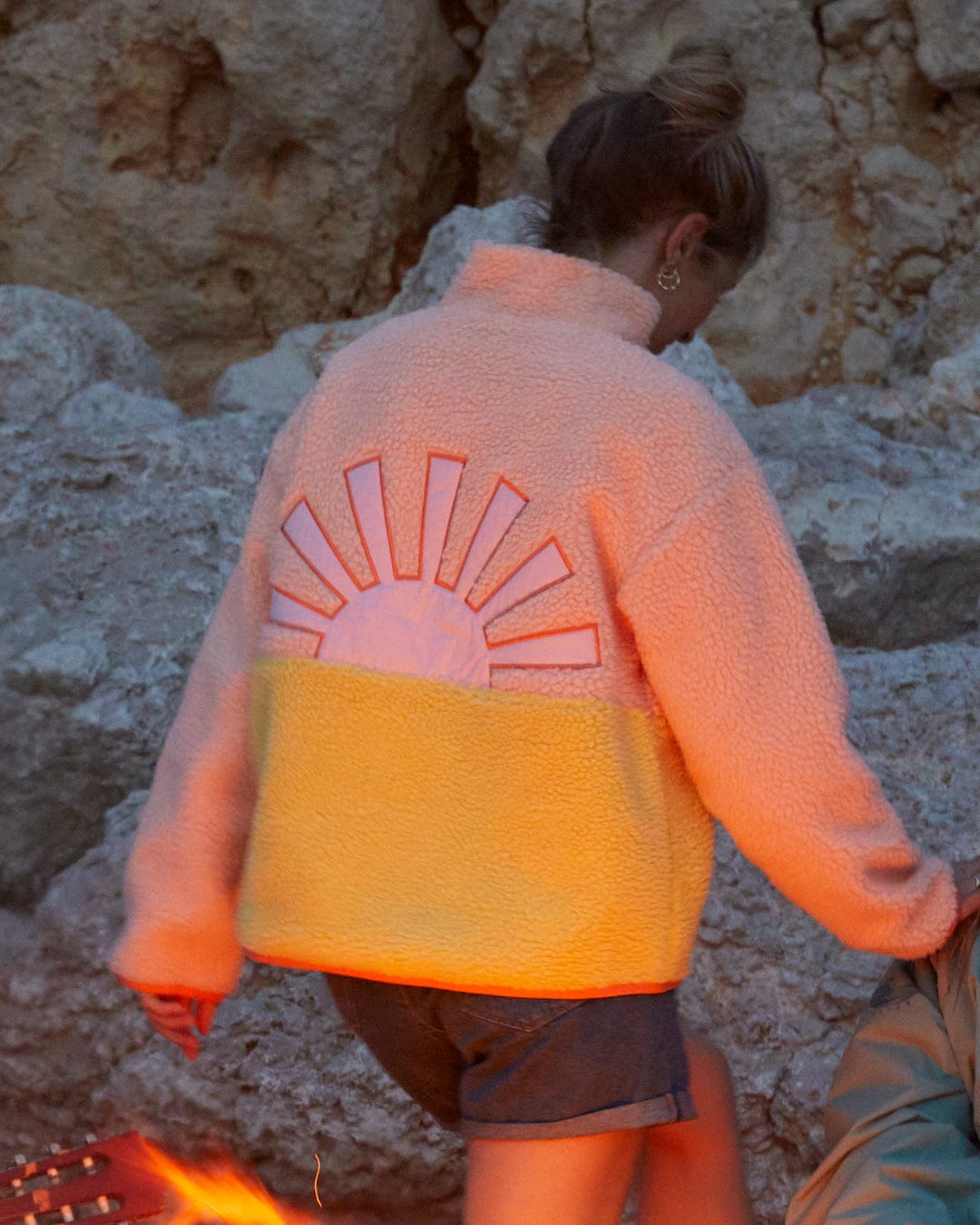 A woman is standing next to a campfire wearing an Emery Sunshine - Womens Sherpa Fleece - Coral jacket by Saltrock with a sun graphic on it.