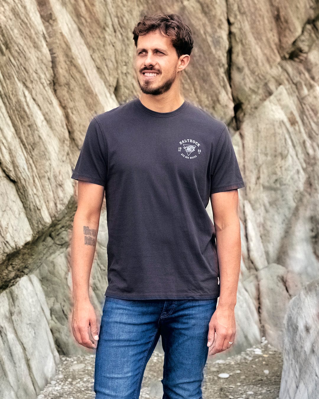 A man wearing a black Eye Sea Waves - Mens Short Sleeve Stonewash T-shirt - Dark Grey and jeans standing next to rocks featuring Saltrock branding.