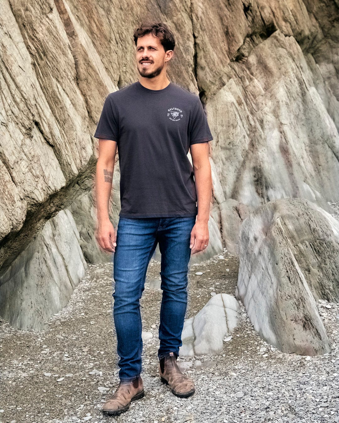 A man wearing an Eye Sea Waves - Mens Short Sleeve Stonewash T-shirt - Dark Grey standing in front of rocks representing the Saltrock branding.
