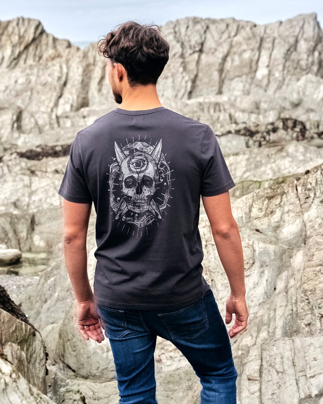 The back of a man wearing a Saltrock branded Eye Sea Waves - Mens Short Sleeve Stonewash T-shirt - Dark Grey with a skull on it.