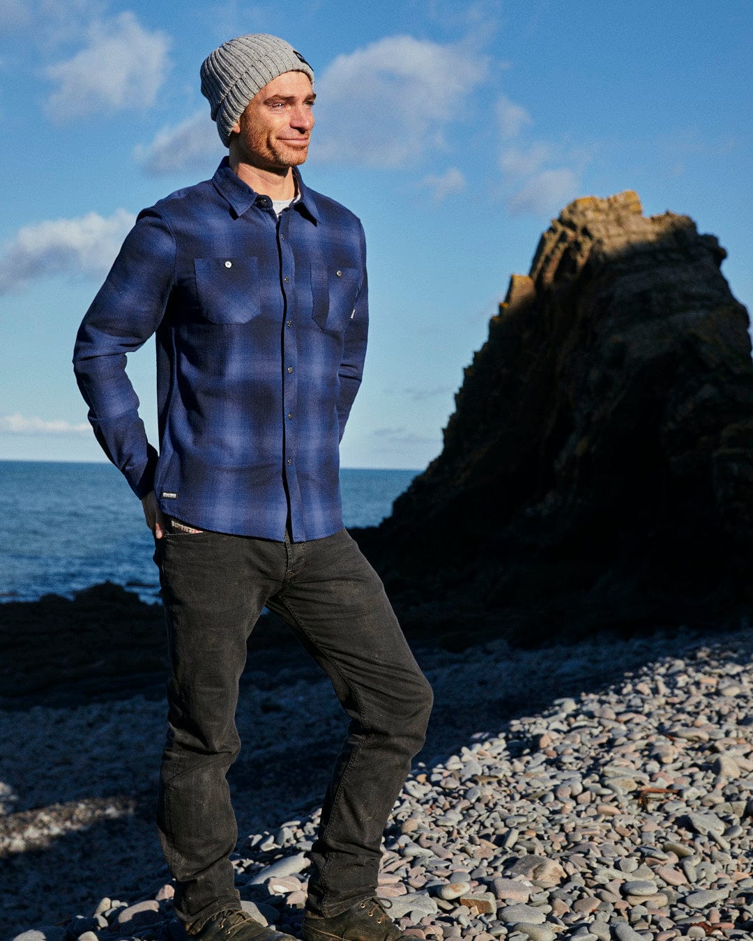 A man standing on a rocky beach with a Saltrock Farris - Mens Check Shirt - Blue beanie on his head.
