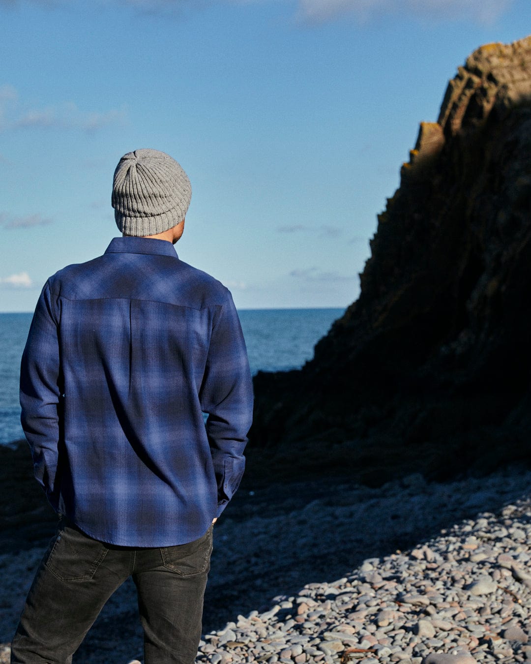 A man standing on a rocky beach, wearing Saltrock's Farris - Mens Check Shirt - Blue made of 100% cotton, gazing out at the vast ocean spread before him under the clear blue sky.