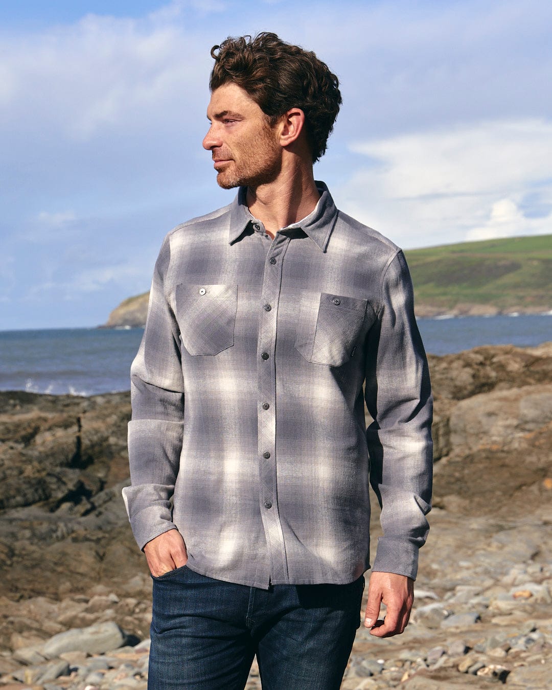 A man wearing a Saltrock Farris - Mens Long Sleeve Check Shirt - Grey, crafted with quality materials, standing on a rocky beach.