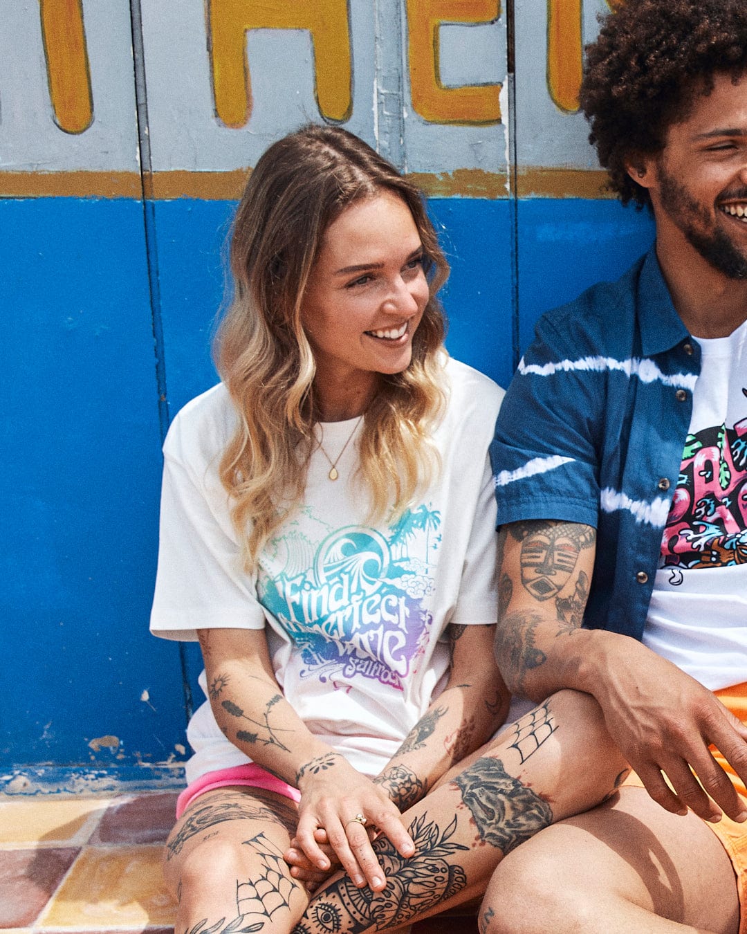 A young man and woman with tattoos sitting and laughing in front of a multi-coloured graffiti wall, wearing the Find The Perfect Wave - Womens Short Sleeve T-Shirt by Saltrock.