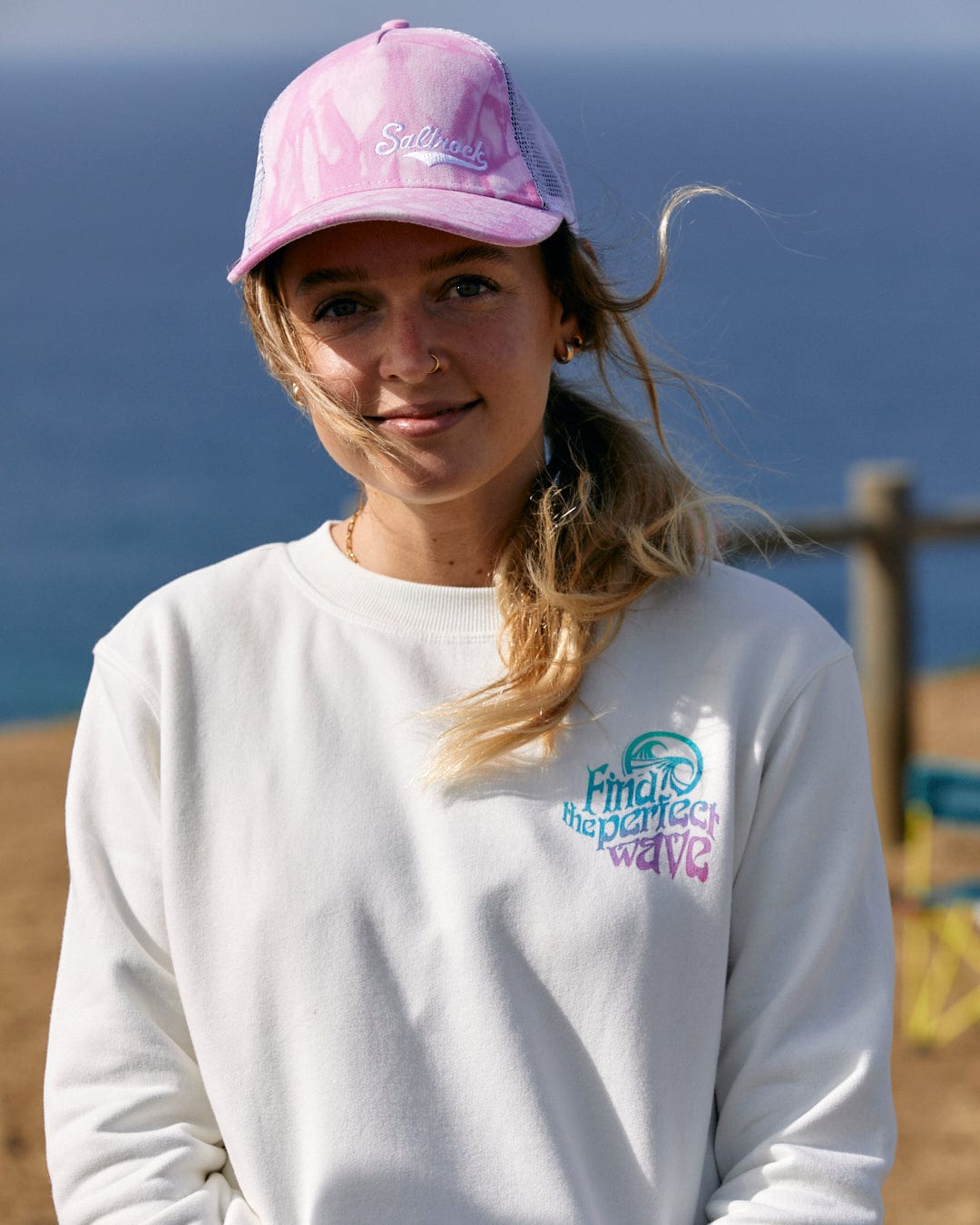 A woman wearing a Saltrock Find The Perfect Wave - Womens Sweat - White with a multi-coloured slogan and a pink hat.