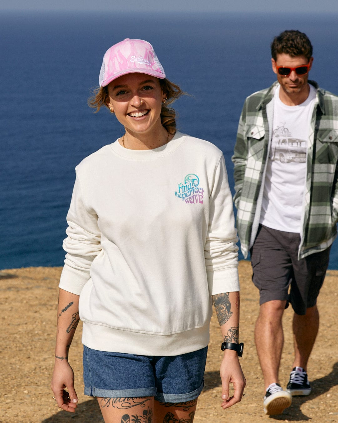 A man and woman walking down a hill next to the ocean, both wearing Saltrock 100% Cotton tees with a multi-coloured slogan.