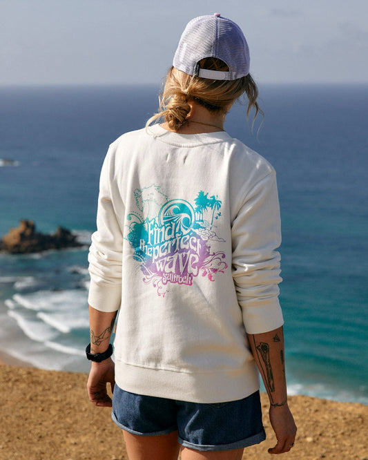 A woman wearing a white Saltrock Find The Perfect Wave - Womens Sweat sweatshirt with a multi-colored slogan, standing on a hill overlooking the ocean.
