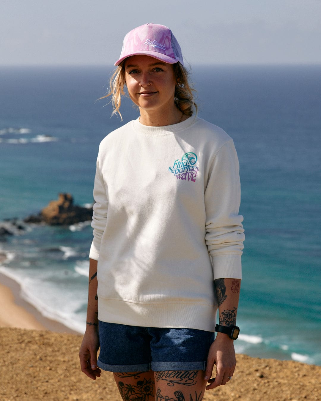 A woman wearing a multi-coloured slogan hat and Saltrock Find The Perfect Wave - Womens Sweat - White standing on a hill near the ocean.