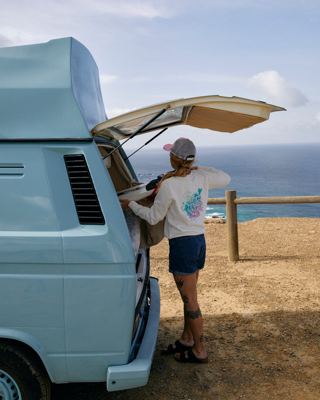 Person preparing food at the back of a campervan with a scenic coastal backdrop, wearing a Saltrock Find The Perfect Wave - Womens Sweat - White crew neck t-shirt.