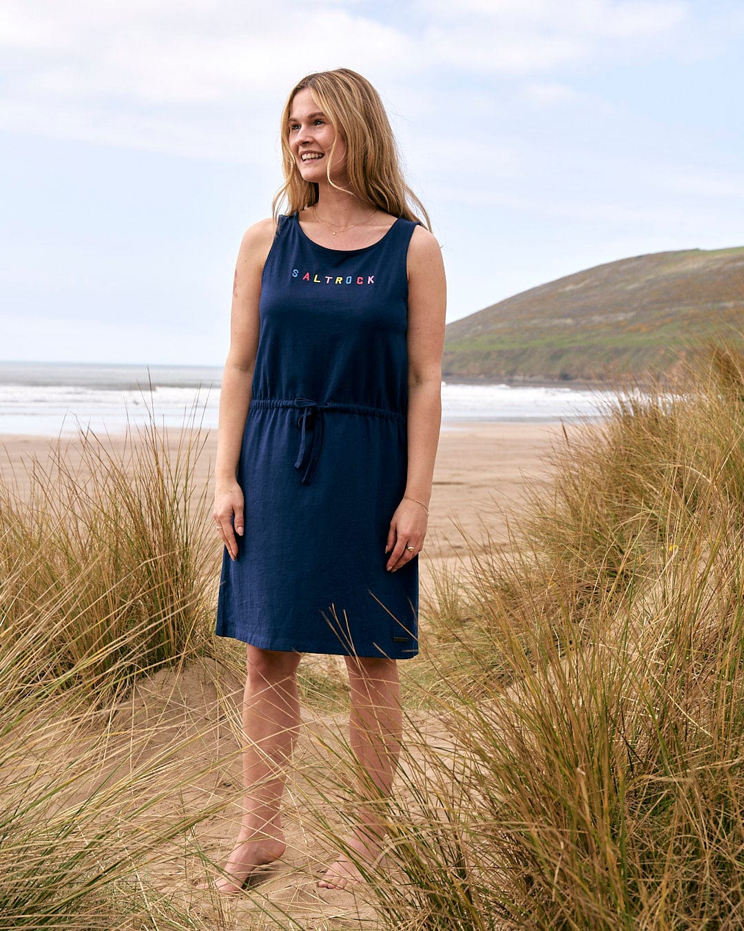 A woman standing on the beach in a Saltrock Flax - Womens Tie Vest Dress - Blue.