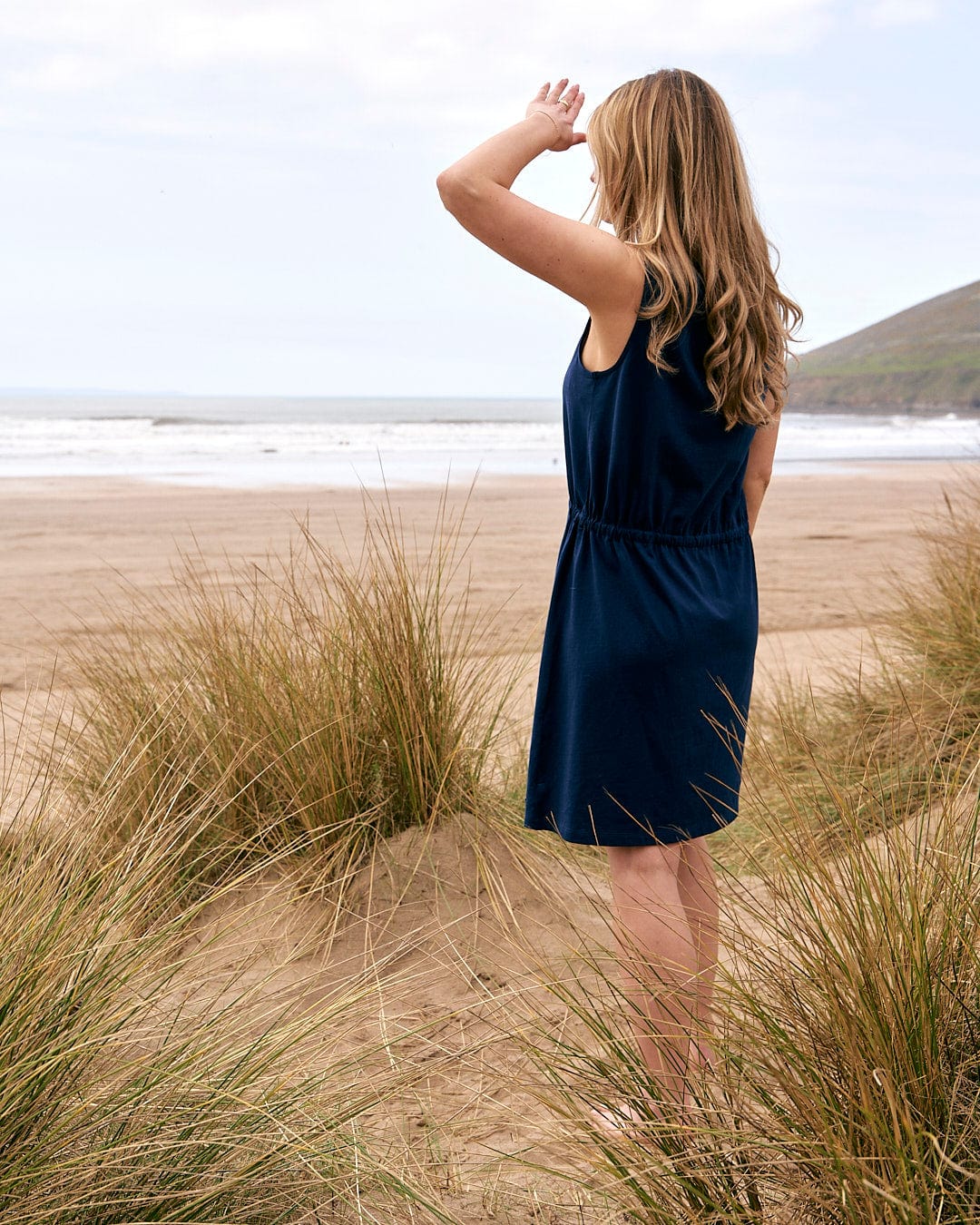 A woman is standing on a beach looking at the ocean wearing the Saltrock Flax - Womens Tie Vest Dress - Blue.
