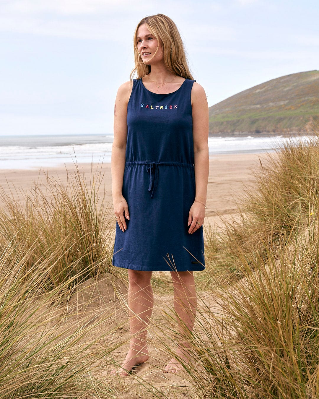 A woman standing on the beach in a Saltrock Flax - Womens Tie Vest Dress - Blue.