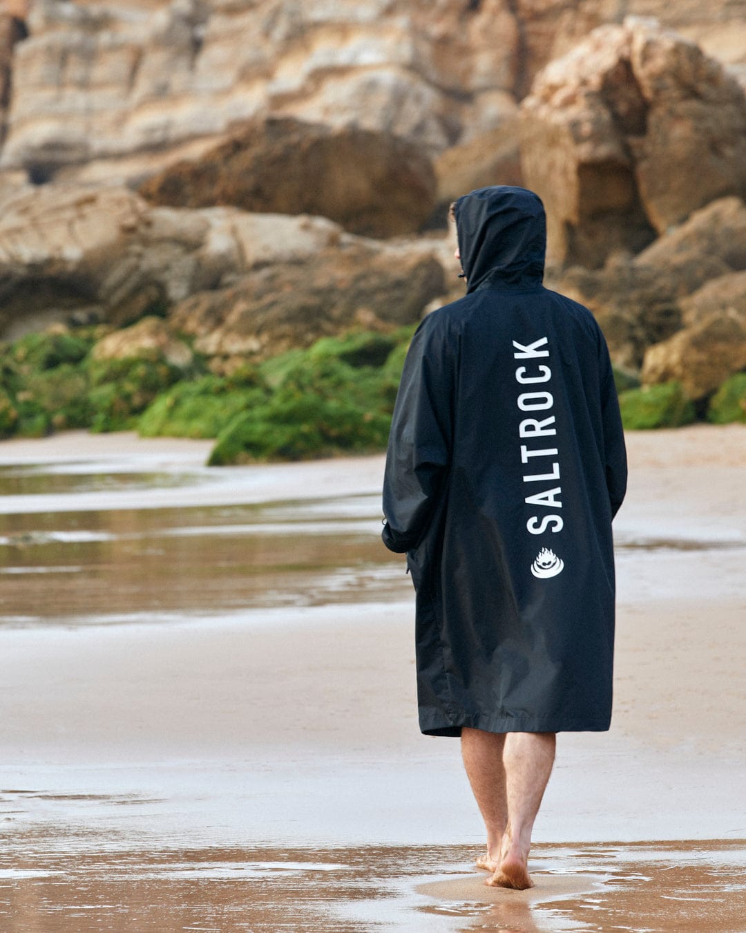 Person wearing a Saltrock 3 in 1 Recycled Four Seasons Changing Robe - Black/Yellow with "Saltrock" logo and towelling lining, standing on a sandy beach facing rocky cliffs.