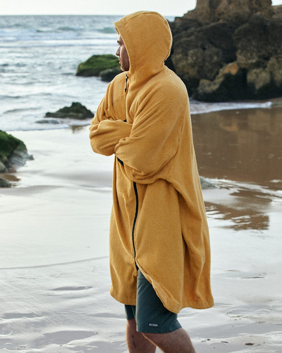 Man in a Saltrock 3 in 1 Recycled Four Seasons Changing Robe - Black/Yellow standing on a beach, looking out at the water, with rocks and sand visible around him.