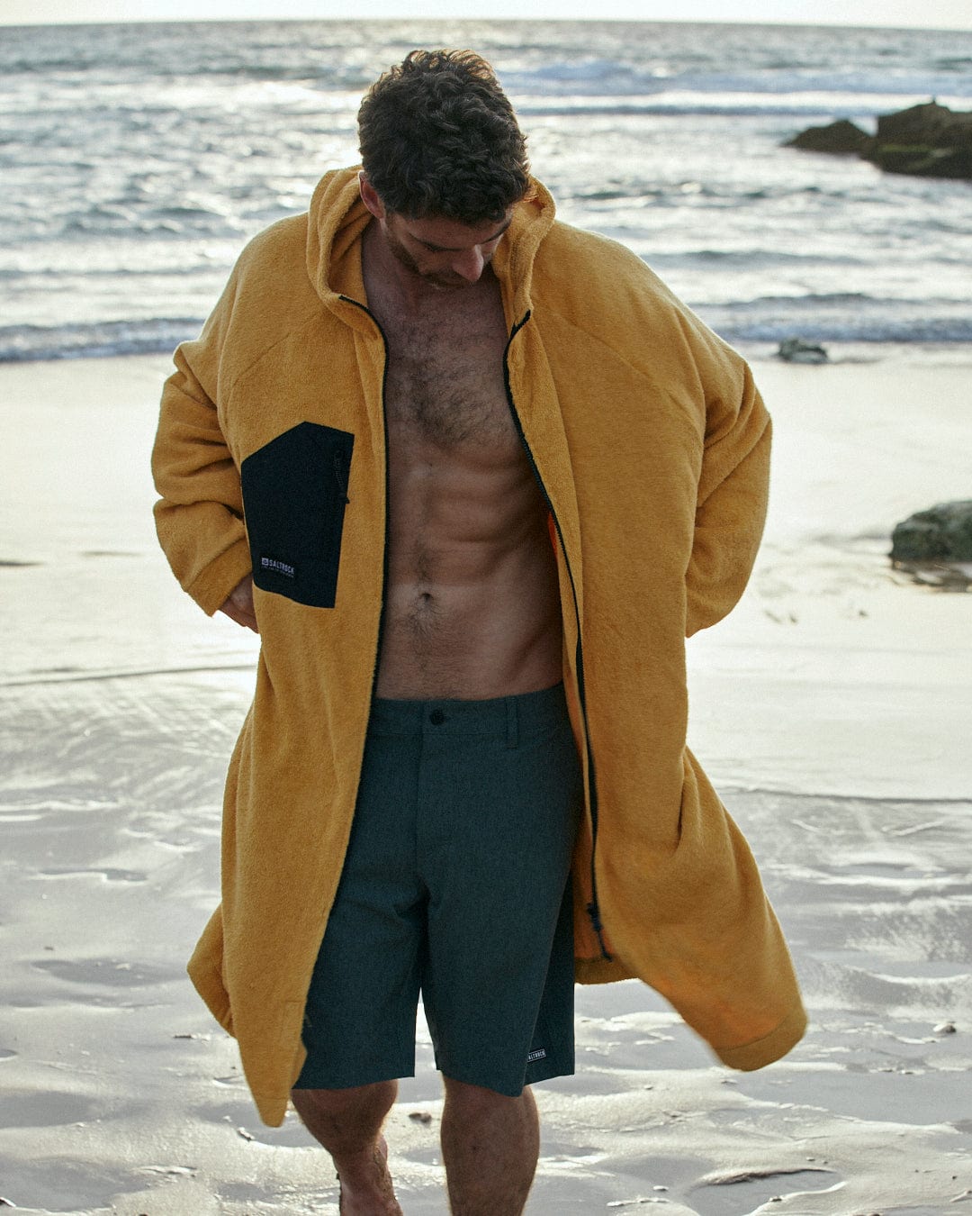 A man in a Saltrock 3 in 1 Recycled Four Seasons Changing Robe - Black/Yellow and navy shorts stands on a beach, looking down with a thoughtful expression.