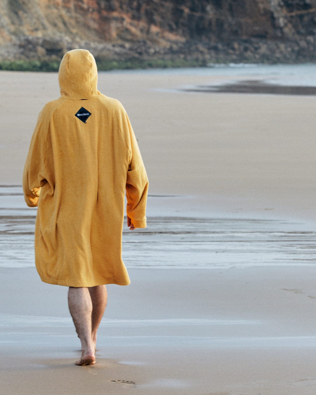 Person in a Saltrock 3 in 1 Recycled Four Seasons Changing Robe - Black/Yellow, walking away from the camera on a sandy beach, with cliffs in the background.