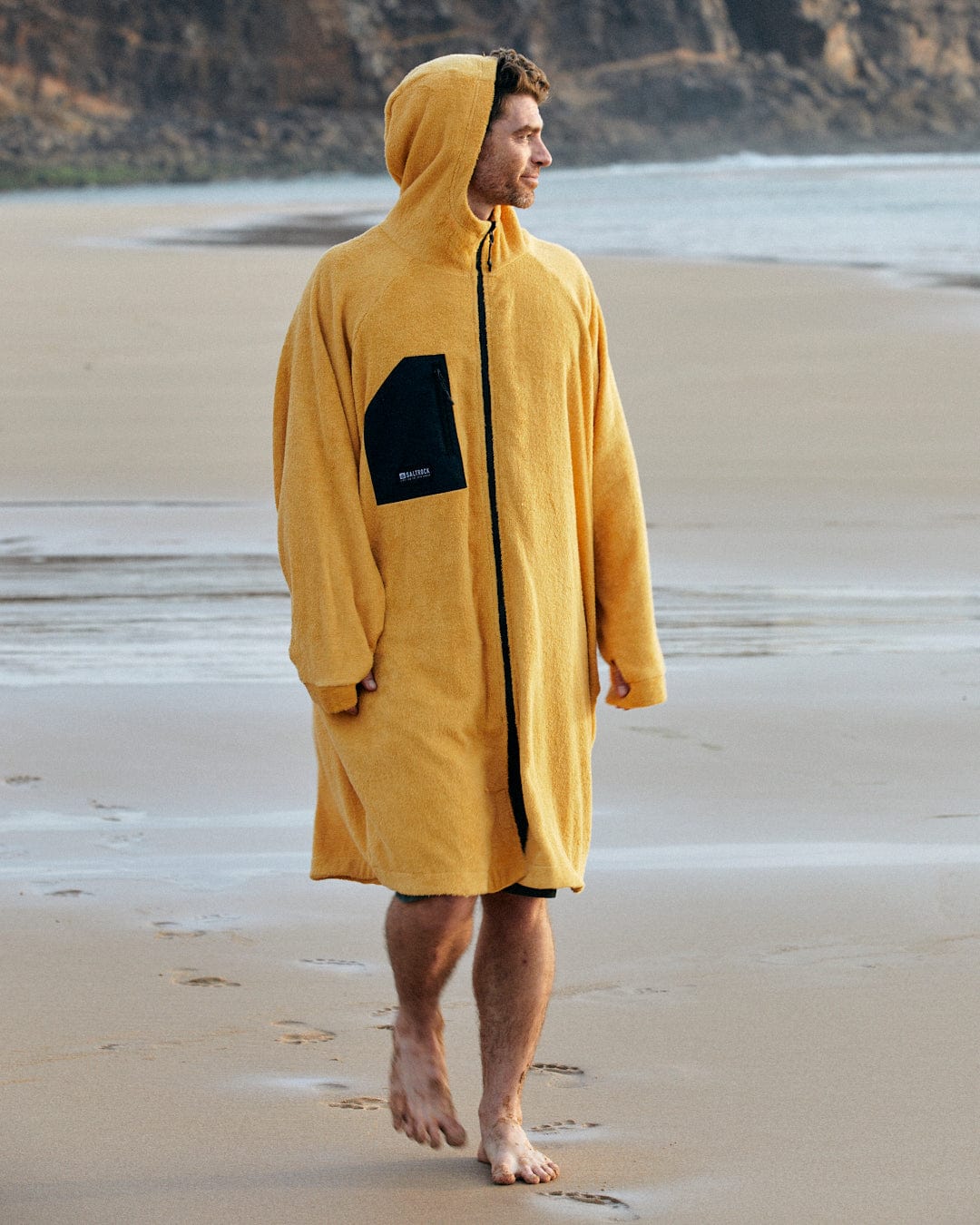 A person wearing a Saltrock 3 in 1 Recycled Changing Robe - Black/Yellow walks barefoot on a sandy beach with rocky cliffs in the background.