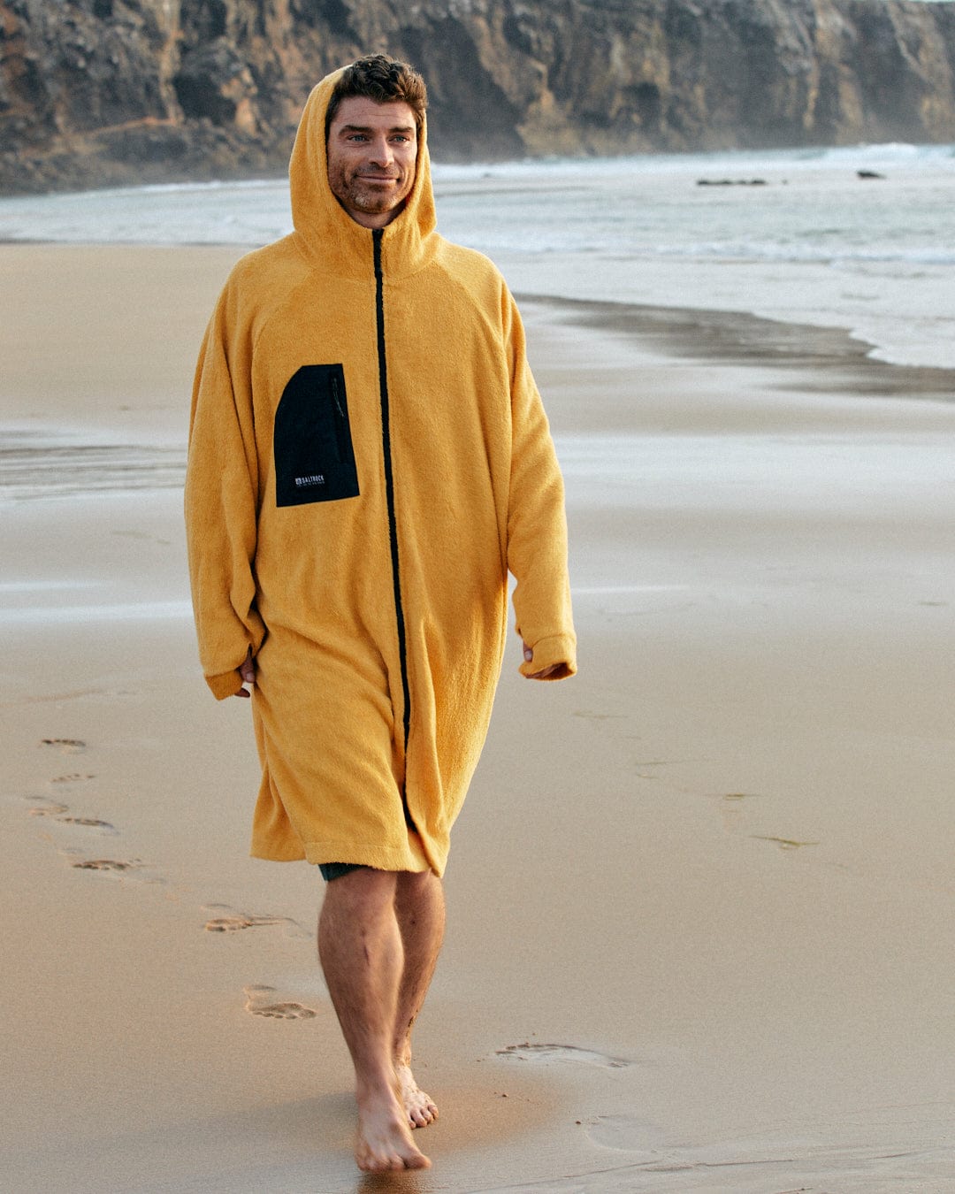 Man in a Saltrock 3 in 1 Recycled Four Seasons Changing Robe - Black/Yellow walking on a sandy beach with ocean waves in the background.