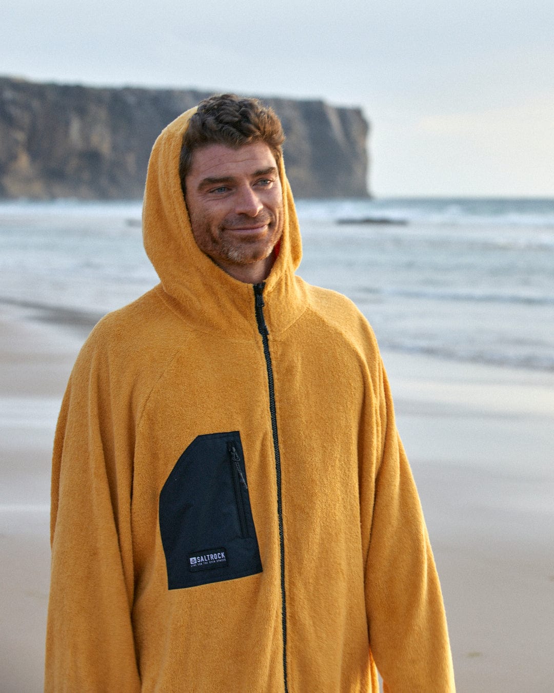 Man in a yellow Saltrock 3 in 1 Recycled Four Seasons Changing Robe smiling on a sandy beach, with a cliff in the background.