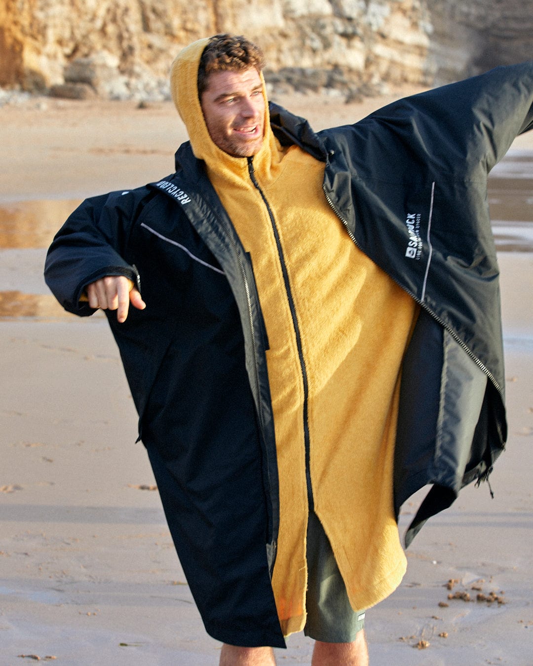 A man in a Saltrock 3 in 1 Recycled Four Seasons Changing Robe - Black/Yellow smiles and spreads his arms on a sandy beach.