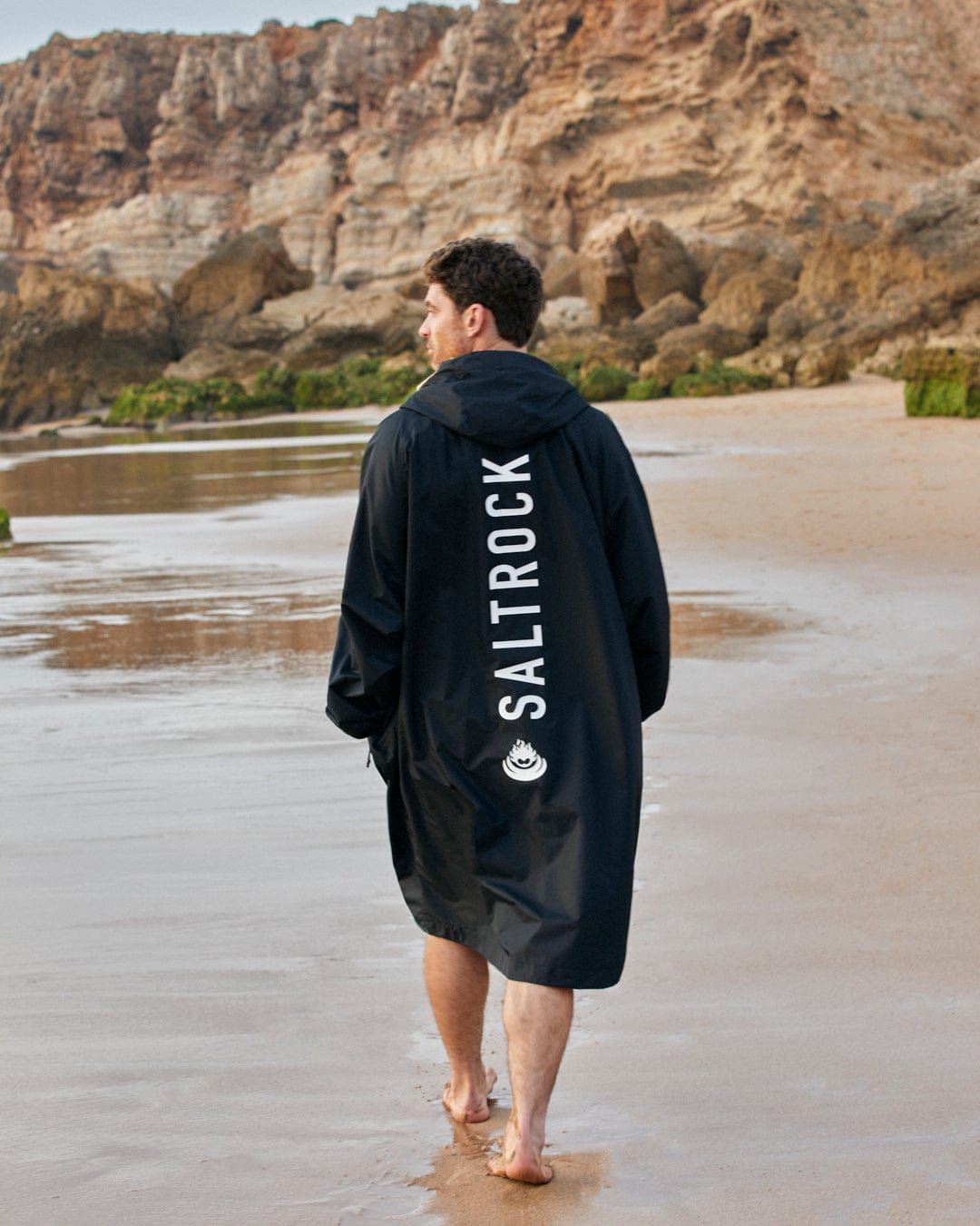 Man in a Saltrock-branded 3 in 1 Recycled Four Seasons Changing Robe - Black/Yellow walking on a sandy beach with rocky cliffs in the background.