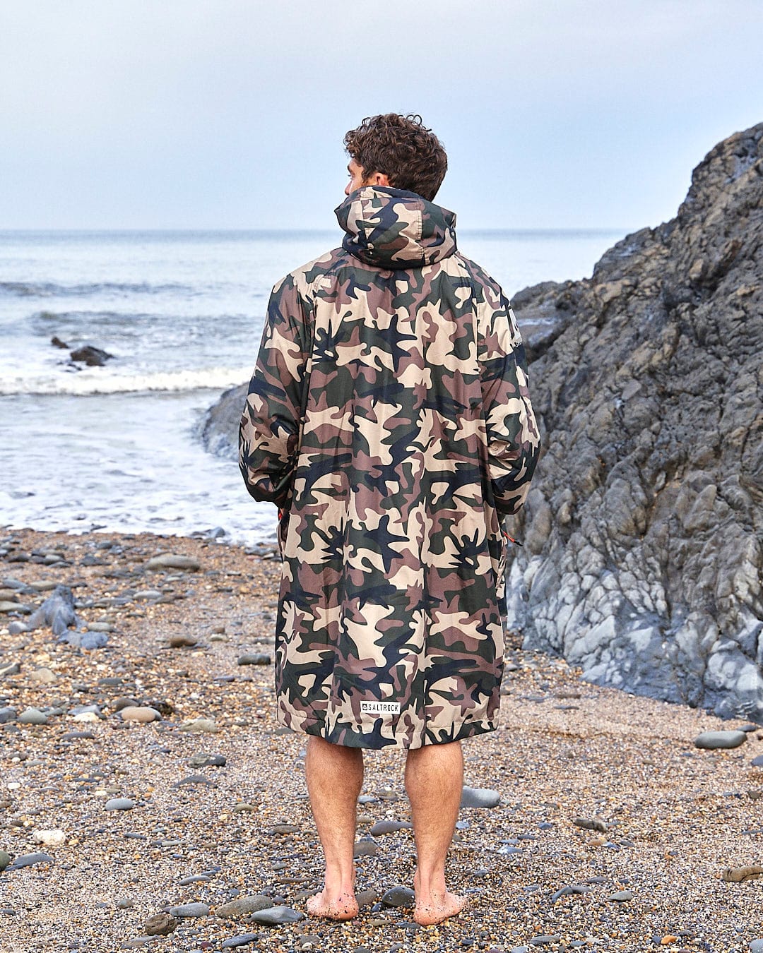 A person stands on a pebble beach, facing the sea, wearing a Saltrock Recycled Four Seasons Changing Robe in Brown Camo with a hood.