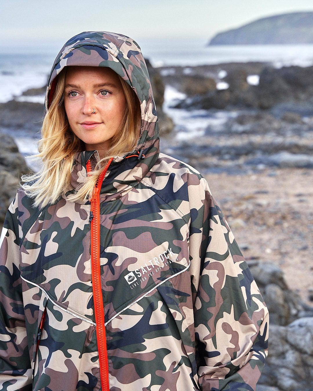 Woman in a Saltrock Recycled Four Seasons Changing Robe - Brown Camo, standing on a rocky beach with the ocean and cliffs in the background.